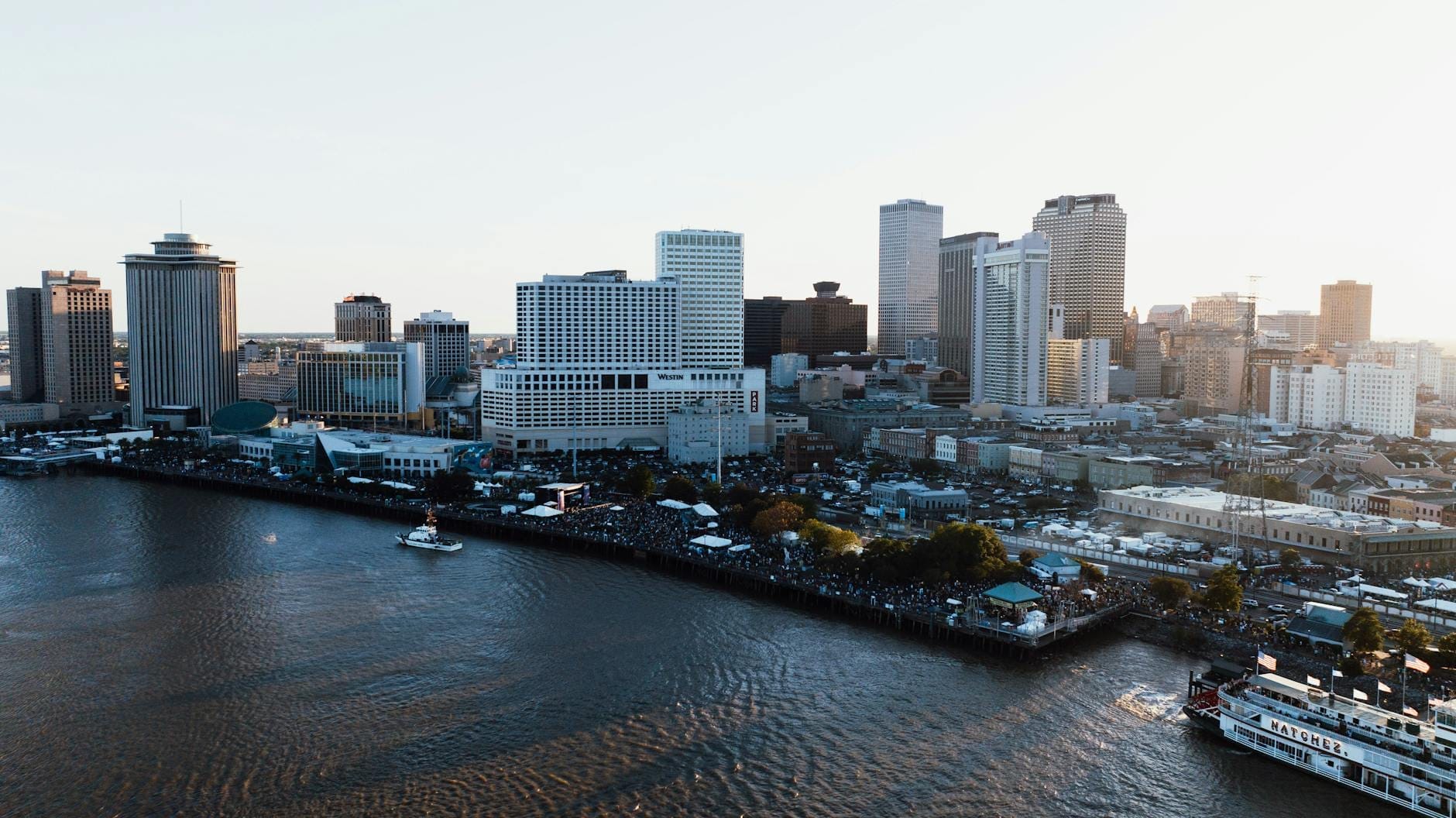 Vista panorâmica do centro de Nova Orleans com o Rio Mississippi em destaque, arranha-céus e barcos no rio sob um céu claro.