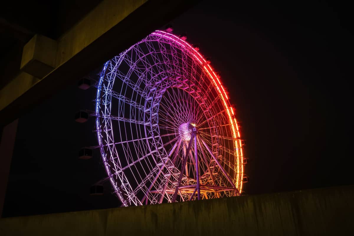 La noria de observación ICON Park iluminada de noche en Orlando, con luces de colores del arcoíris, parcialmente enmarcada por un paso elevado.