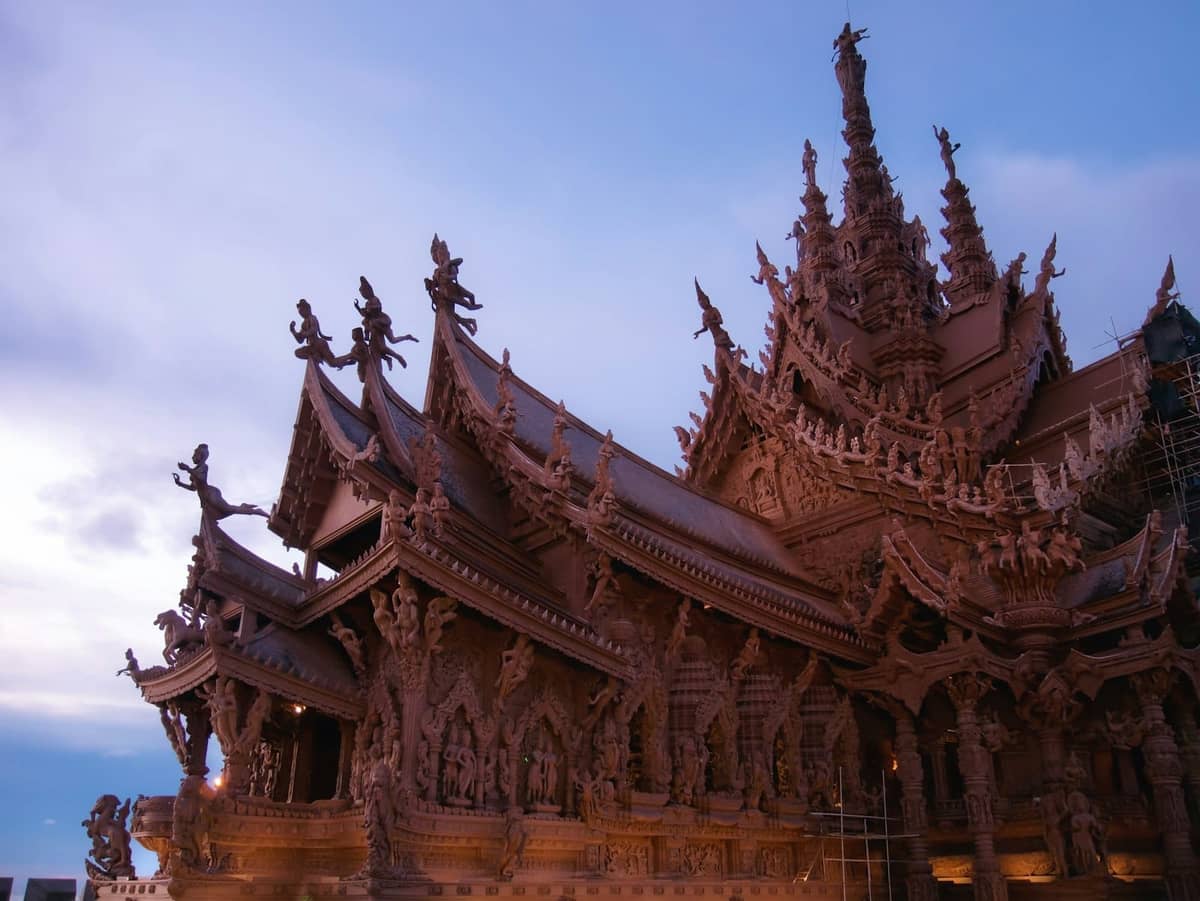Wide angle view of the Sanctuary of Truth temple in Pattaya at dusk, featuring elaborate wooden carvings and intricate roof details.