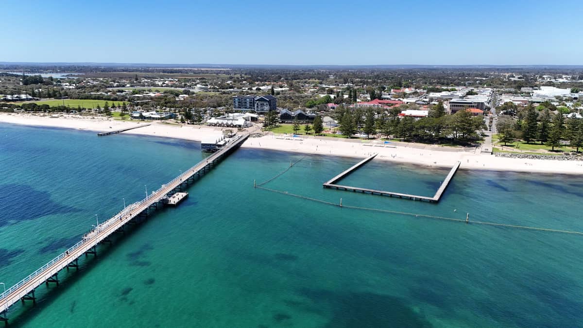 Luftaufnahme eines Sandstrands in einer städtischen Umgebung mit einem langen Pier, ruhigem türkisfarbenem Ozean und abgesperrtem Schwimmbereich.