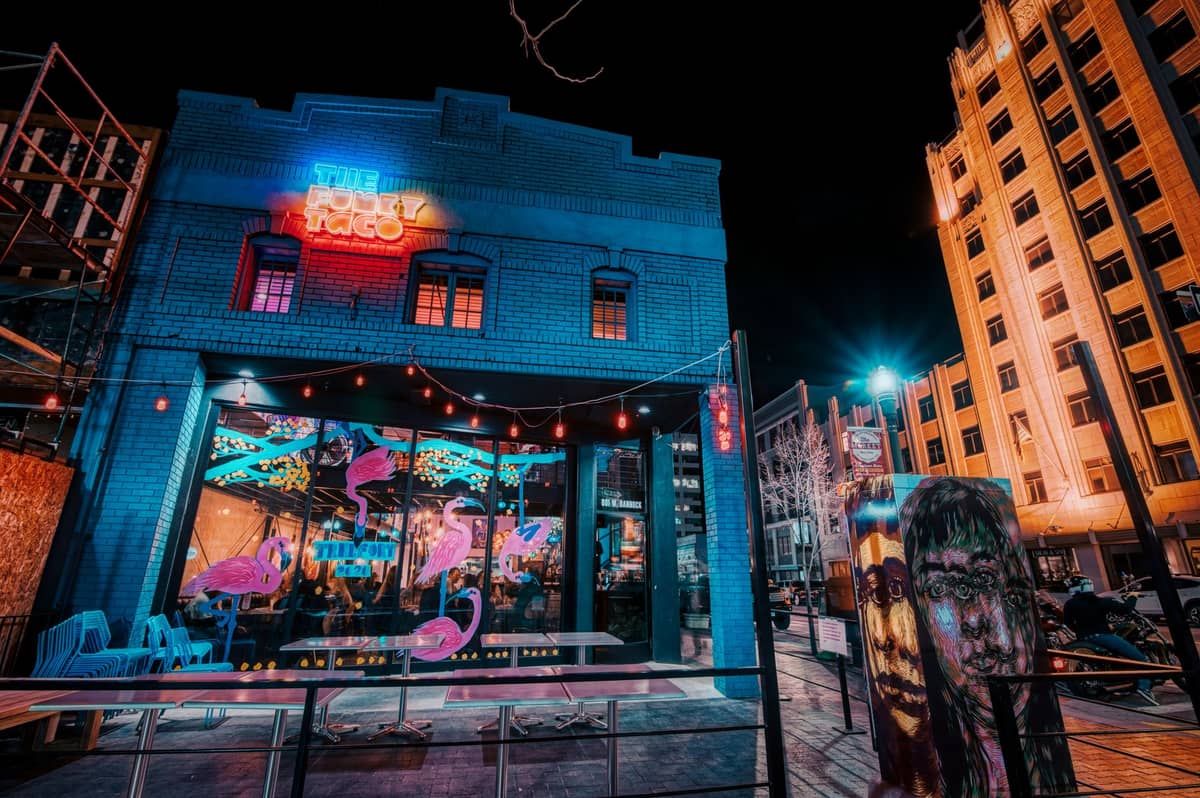 Ein lebhaftes, modernes Restaurant mit Neon-Schildern und Gästen im Inneren an einer Stadtstraße bei Nacht, umgeben von hohen Gebäuden.