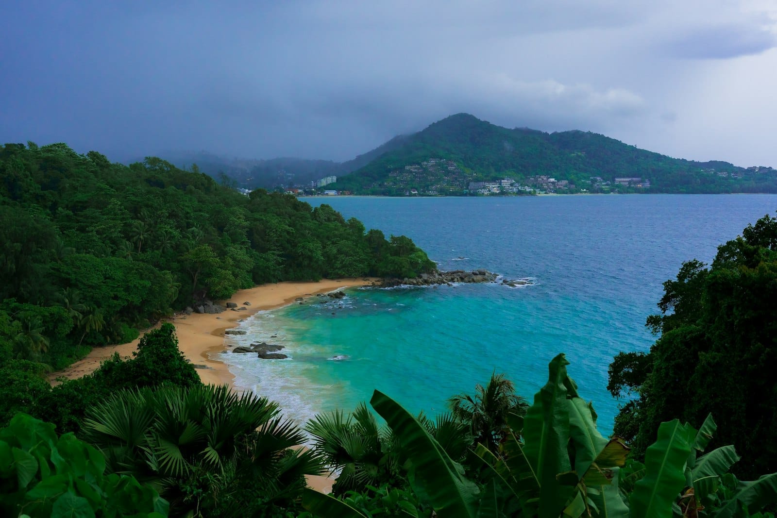 Secluded sandy beach with turquoise water, lush green hills, and distant resorts, showing a quieter side of Phuket beyond busy Patong.