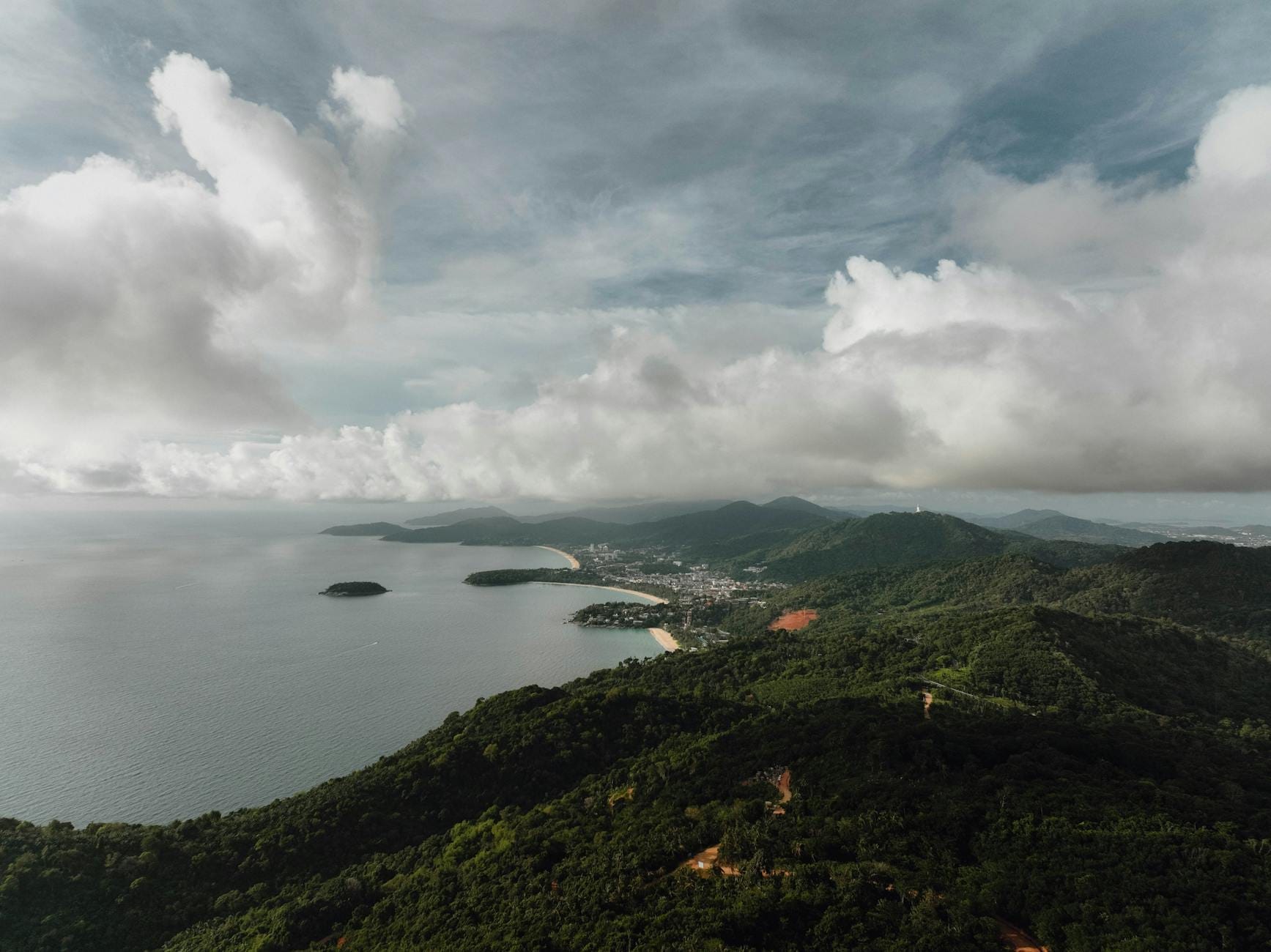 A sweeping aerial view of Phuket's forested hills, beaches, and coastline under a dramatic cloudy sky, showcasing the island's natural landscape.