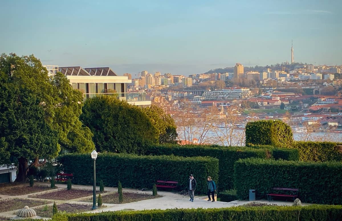 Persone che camminano lungo sentieri di giardino curati con alte siepi e panchine, con vista sul fiume Douro e il paesaggio urbano di Porto sullo sfondo.