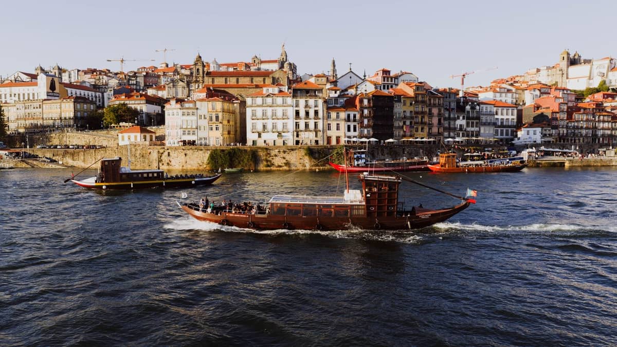 Tradizionali battelli da crociera sul fiume Douro che passano davanti a variopinti edifici storici sul lungofiume Ribeira di Porto in piena luce diurna.