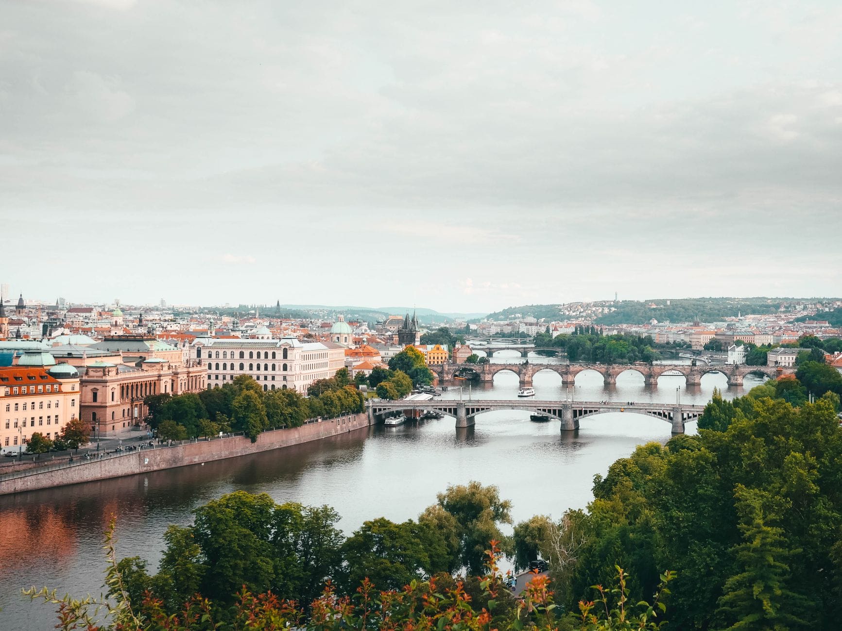 Panoramablick auf Prag mit der Moldau, mehreren historischen Brücken und der malerischen Altstadt, eingerahmt von sattem Grün.