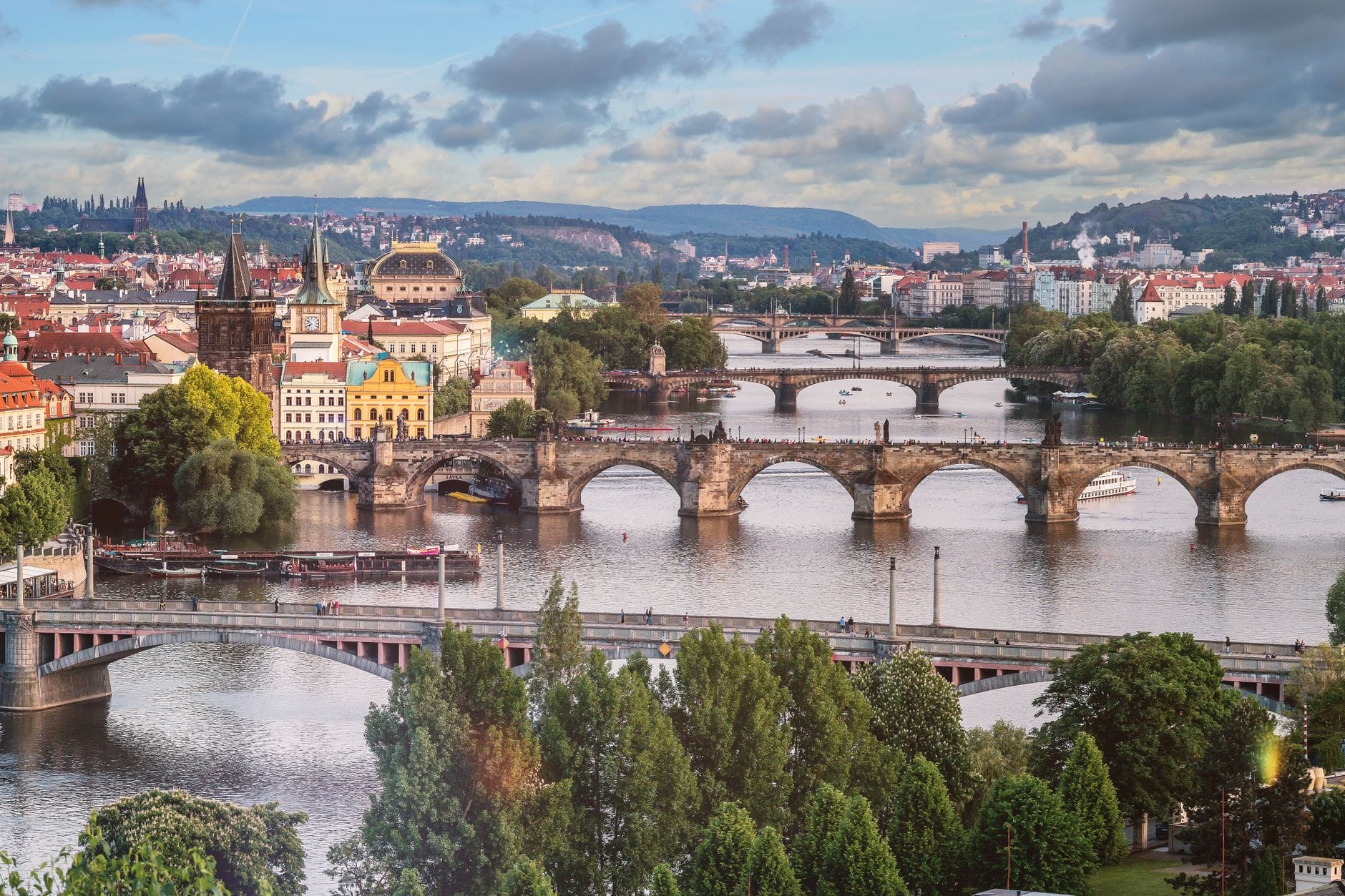 Weites Panorama von Prag mit mehreren Brücken über die Moldau und bunten historischen Gebäuden unter einem bewölkten Himmel.