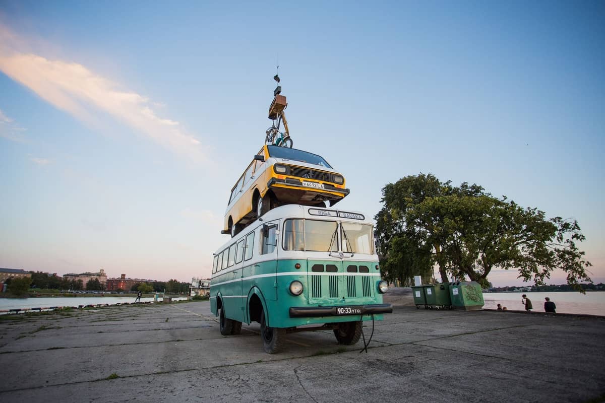Eine skurrile Kunstinstallation am Rigaer Ufer mit einem retrogrünen Bus, auf dem bei Sonnenuntergang ein gelber Van und ein Fahrrad thronen.