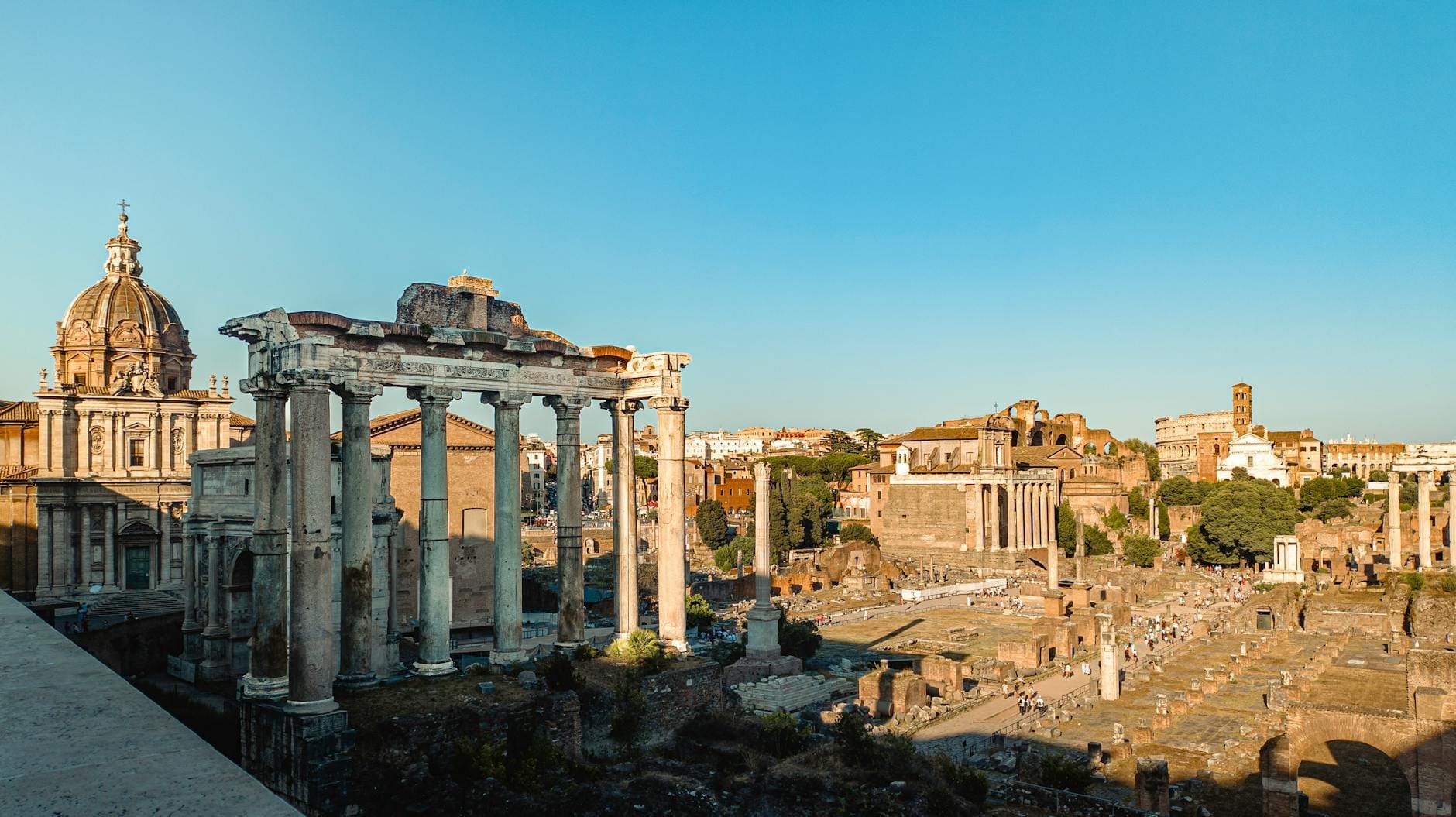 Panoramic view of the Roman Forum with ancient ruins, columns, and surrounding historical buildings under a clear blue sky.