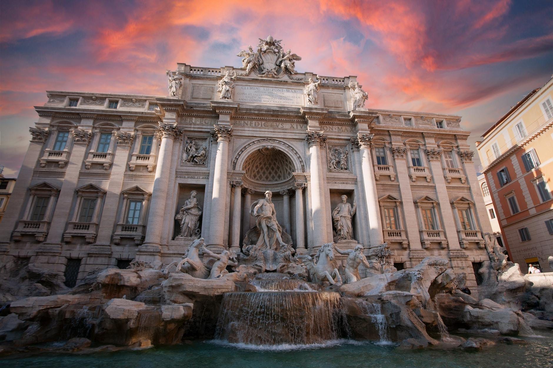 Wide-angle view of Trevi Fountain in Rome at sunset, with detailed sculptures, water cascading, and dramatic colorful sky above the historic architecture.