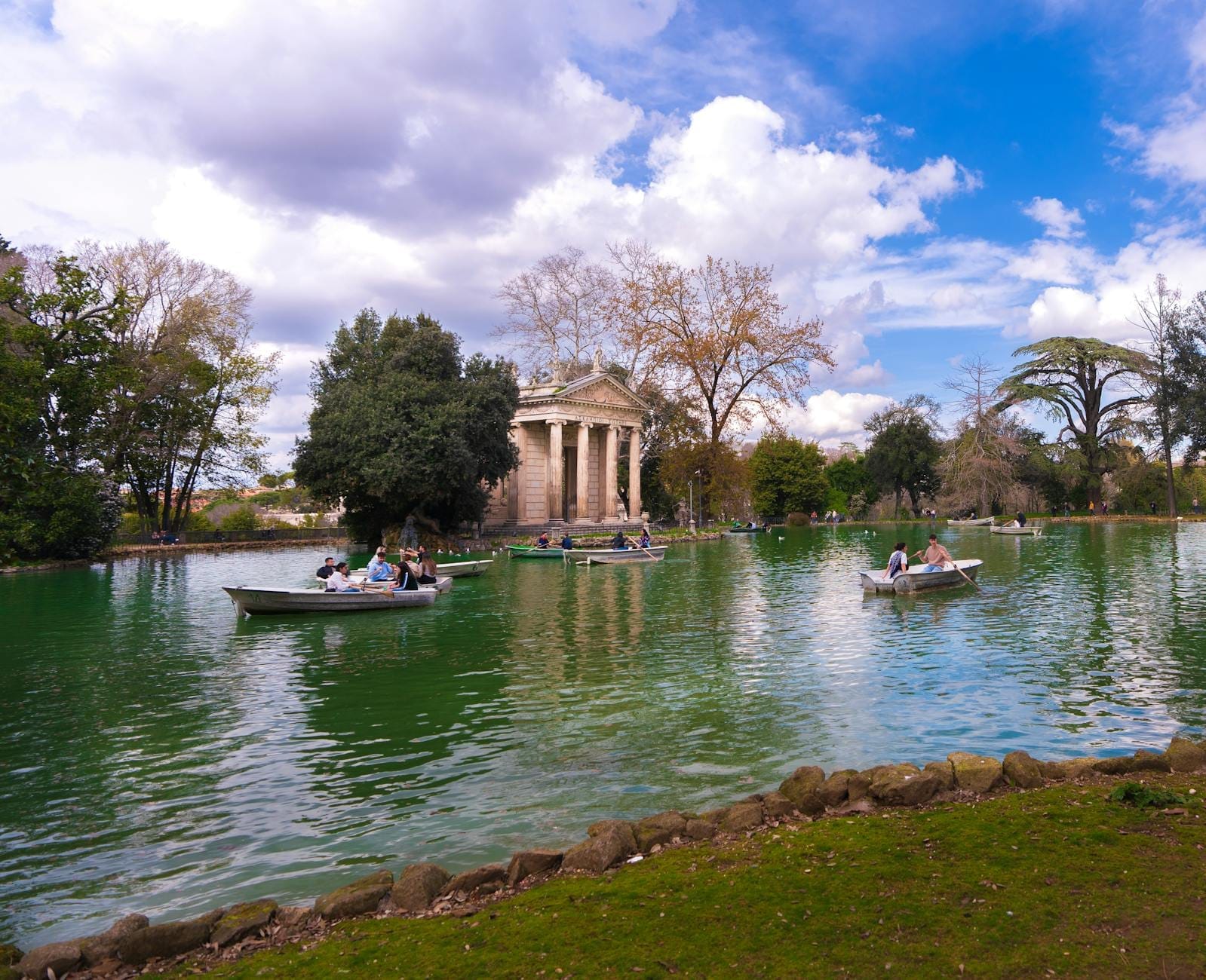 Wide view of the Temple of Aesculapius and lake with rowboats and people inside Rome’s Villa Borghese gardens under a partly cloudy sky.