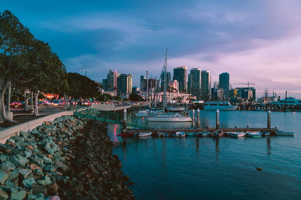 Blick auf die Skyline der Innenstadt von San Diego vom Uferbereich bei Abenddämmerung mit angedockten Booten und Stadtgebäuden im Hintergrund.