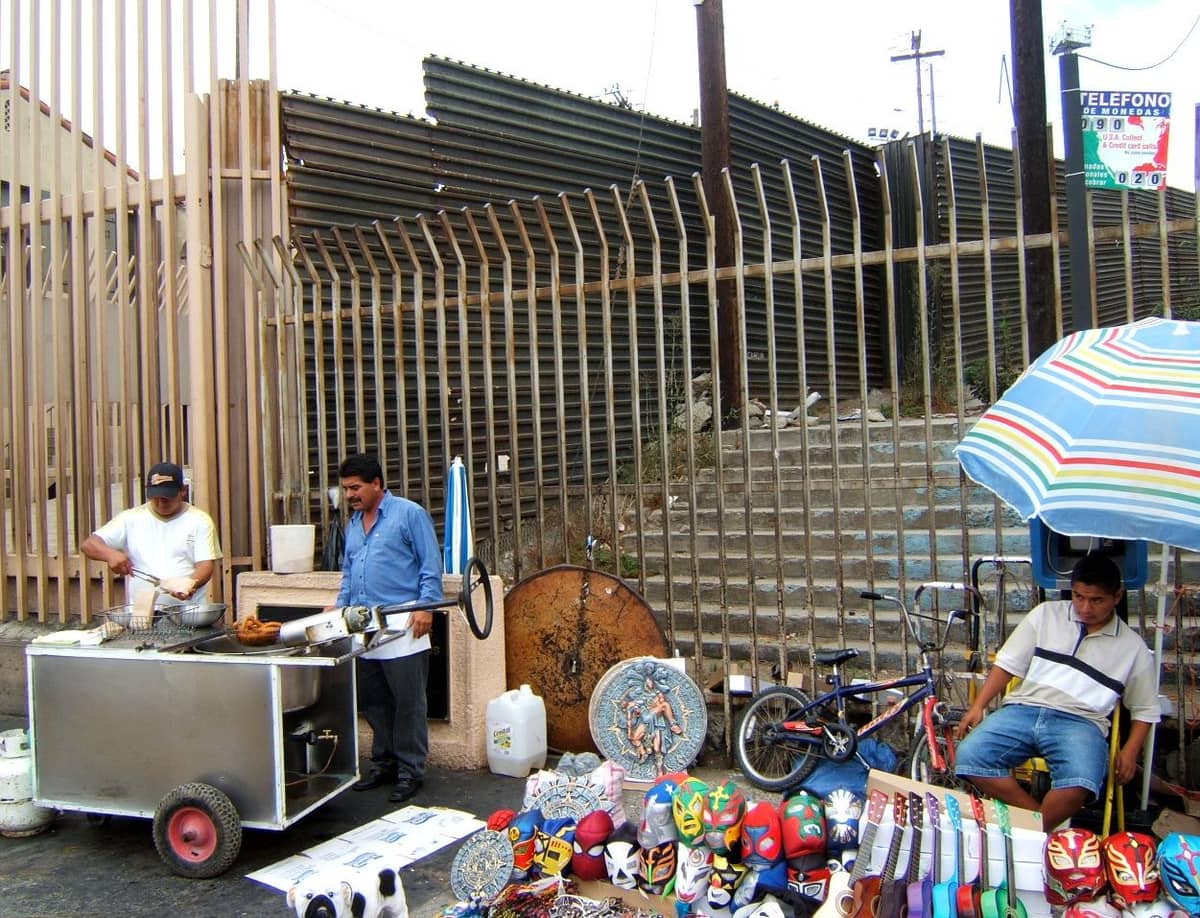Straßenhändler verkaufen Essen und Souvenirs am Grenzzaun zwischen den USA und Mexiko, mit bunten Masken und einem gestreiften Sonnenschirm.