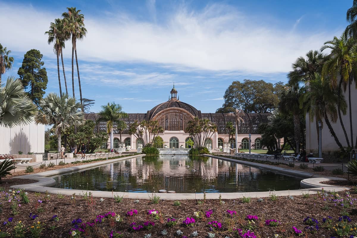 Weitläufige Aussicht auf das Botanical Building und den Seerosenteich des Balboa Park mit Palmen und Blumen unter blauem Himmel in San Diego.