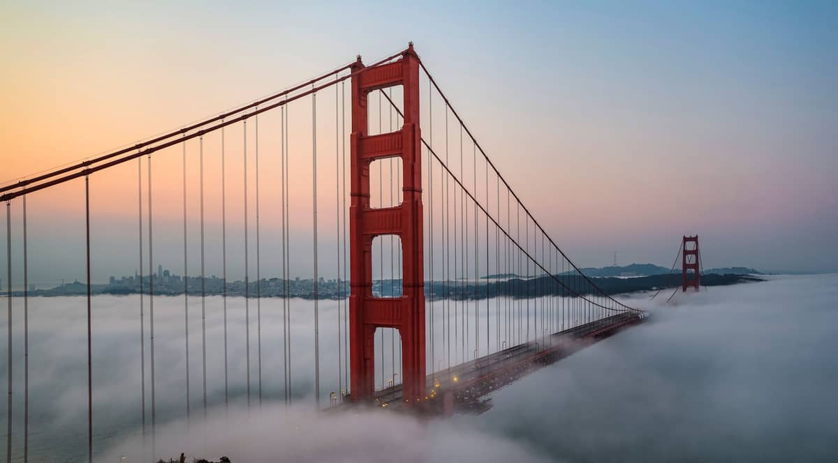 Golden Gate Bridge in San Francisco bei Sonnenaufgang, mit Türmen, die über den dichten Nebel der Bucht ragen, und der Skyline der Stadt im Hintergrund.