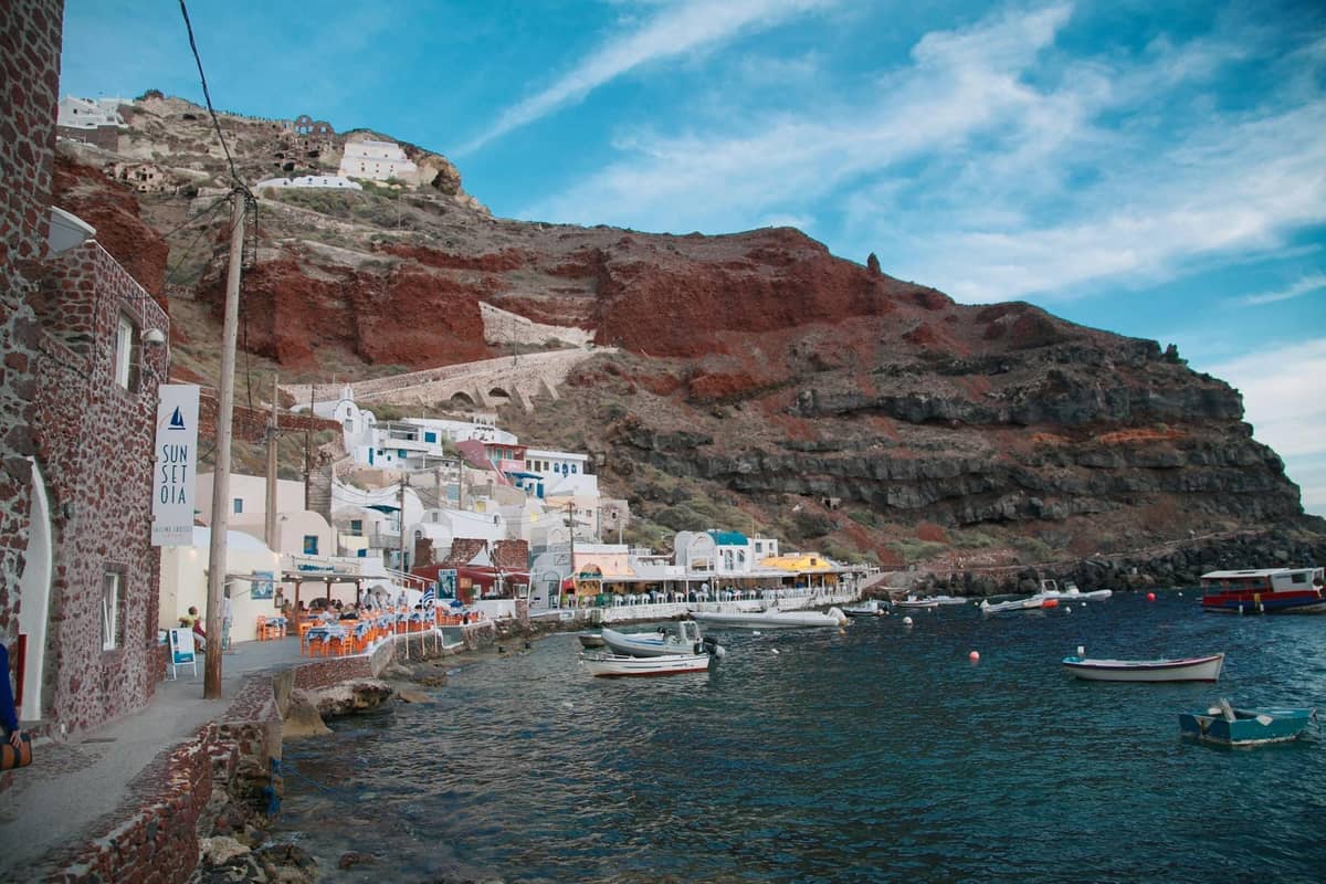 Pequeños botes anclados junto a la orilla de Santorini con acantilados volcánicos y edificios encalados, mostrando la singular costa rocosa.