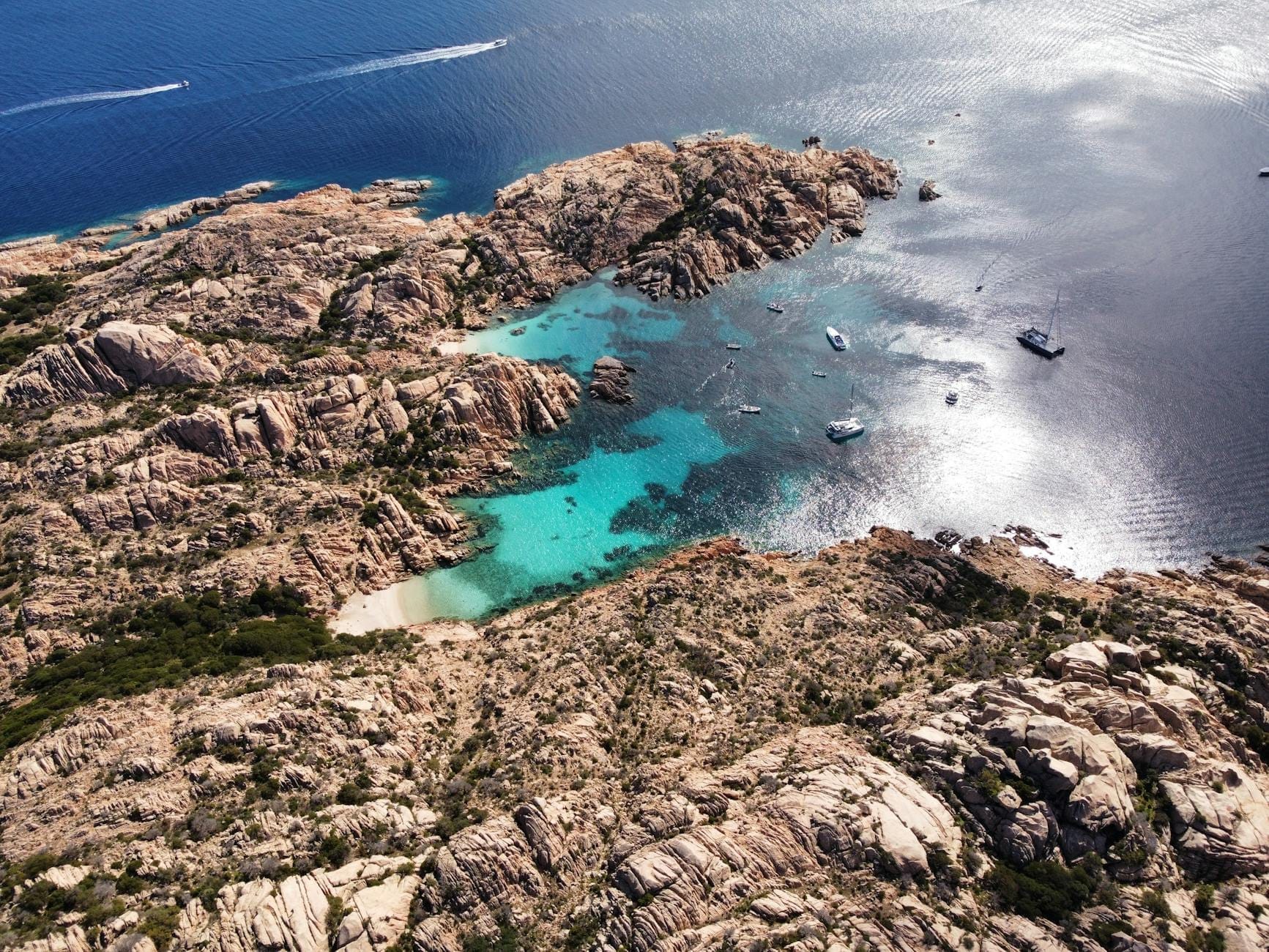 Luftaufnahme einer felsigen Küste mit türkisfarbenen Buchten, kleinen vor Anker liegenden Booten in klarem Wasser und beeindruckender Naturkulisse an der sardischen Küste.