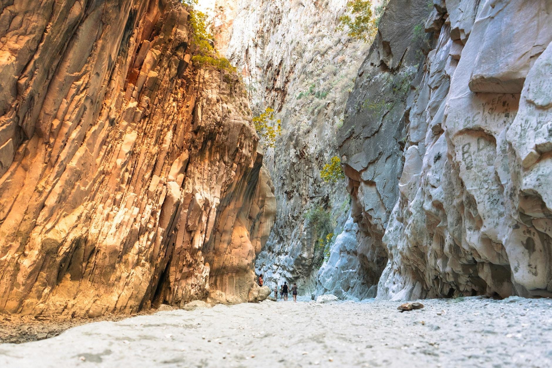 Blick auf eine dramatische Felsschlucht mit sandigem Boden und wandernden Wanderern, die auf ein Outdoor-Abenteuer in der rauen Landschaft Sardiniens hinweist.