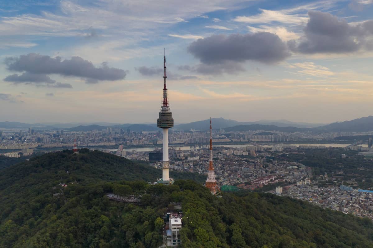 Vista aérea da montanha Namsan e da N Seoul Tower cercadas por uma densa floresta, com o horizonte de Seul e o rio ao fundo sob céu azul.