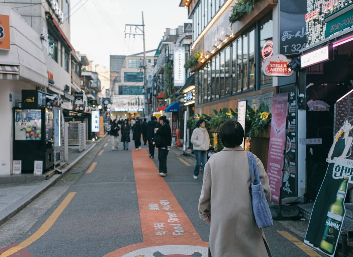 Rua pedonal em Seul repleta de lojas modernas, cafés e pessoas caminhando, refletindo o ambiente animado de um bairro urbano.