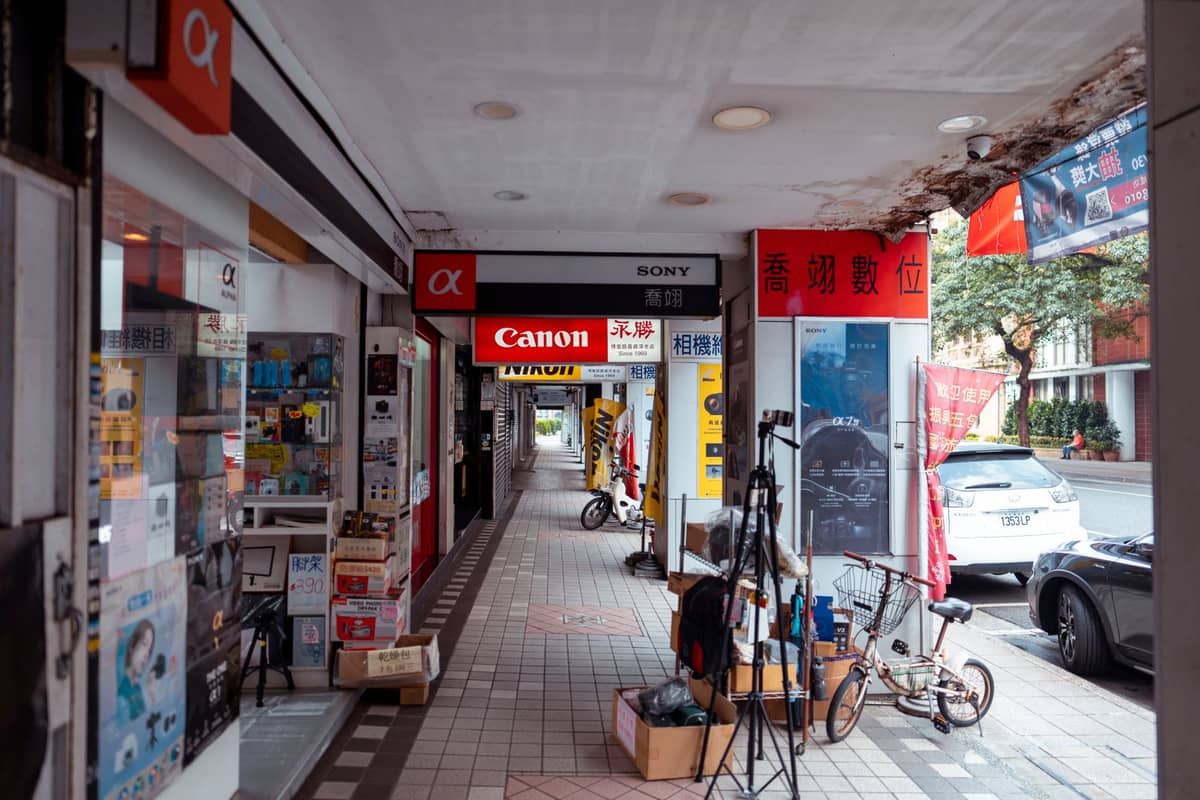 Um corredor ladeado por lojas de eletrônicos em Shenzhen, com marcas como Canon e Sony, e equipamentos e bicicletas na calçada.