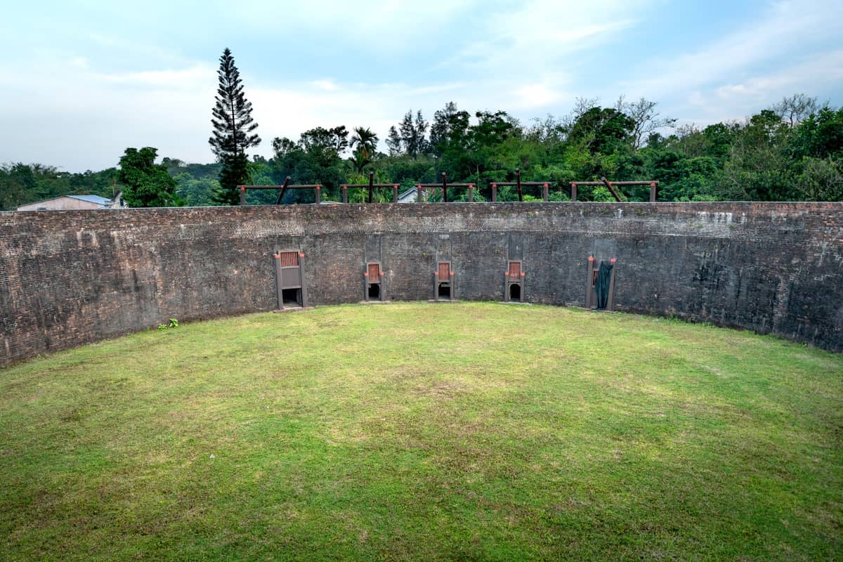 Uma grande fortificação circular de pedra com interior gramado e aberturas de portão originais, rodeada de árvores sob um céu azul no distrito de Longgang.