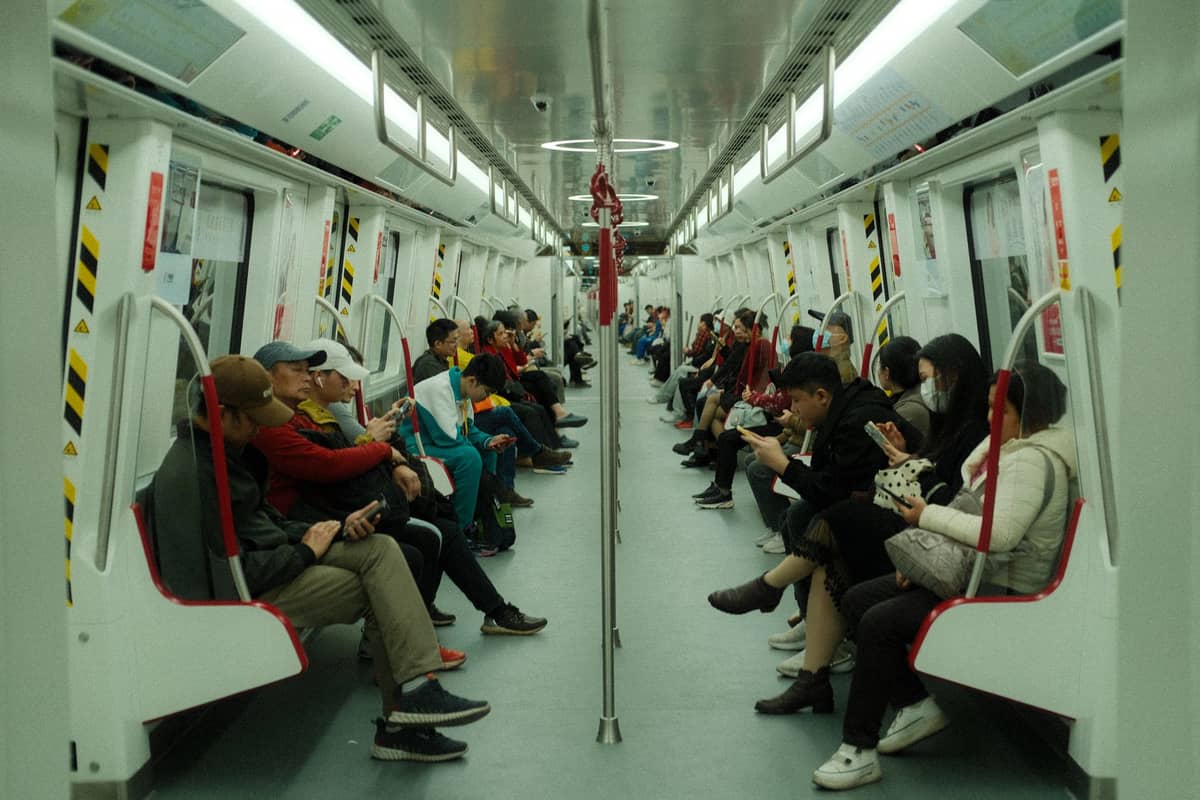 Passageiros sentados dentro de um moderno trem de metrô, com interior branco e corrimãos vermelhos, sugerindo um transporte público eficiente.