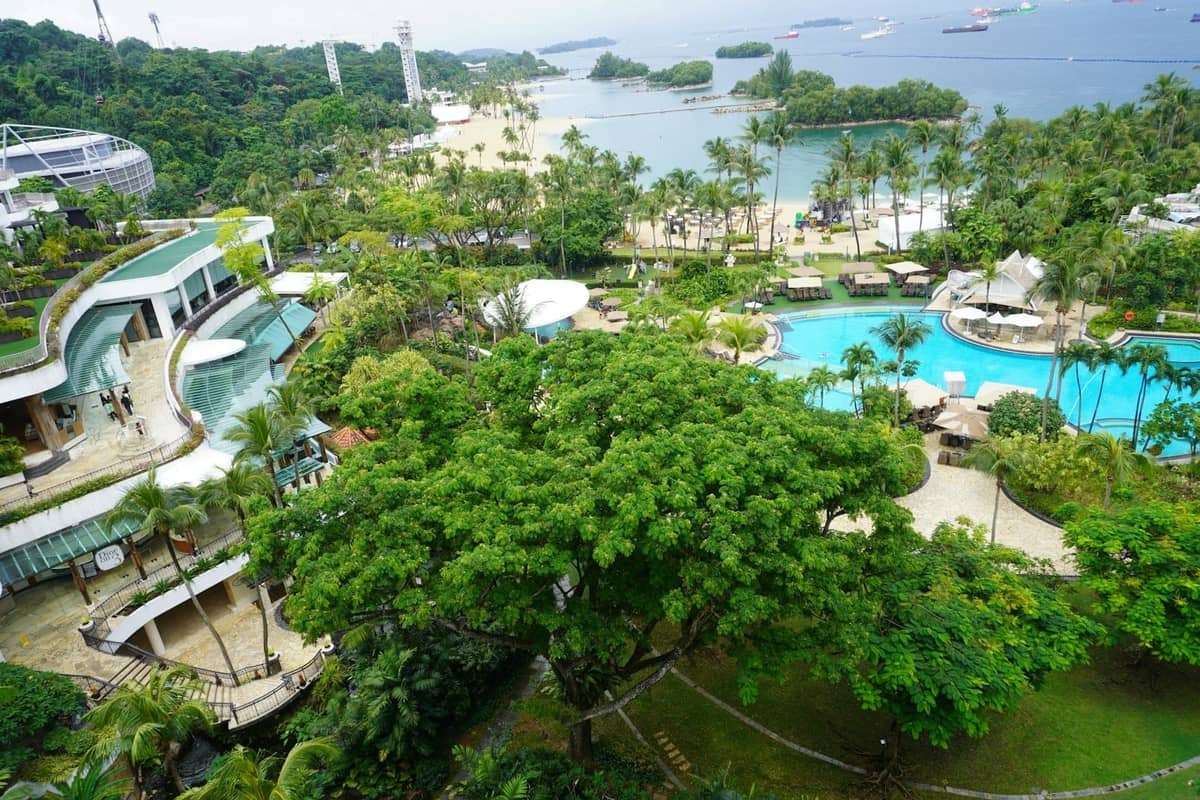 Aerial view of Sentosa Island showing lush greenery, resort pools, beach, and the sea with anchored ships in the background.