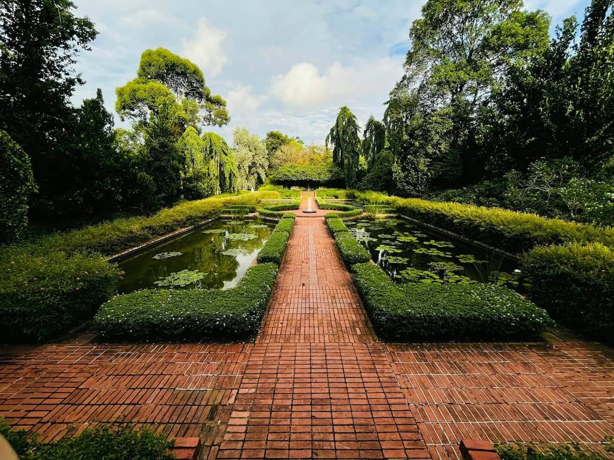 A formal garden in Singapore with a brick pathway, symmetrical ponds, lush green hedges, and trees under a bright sky.
