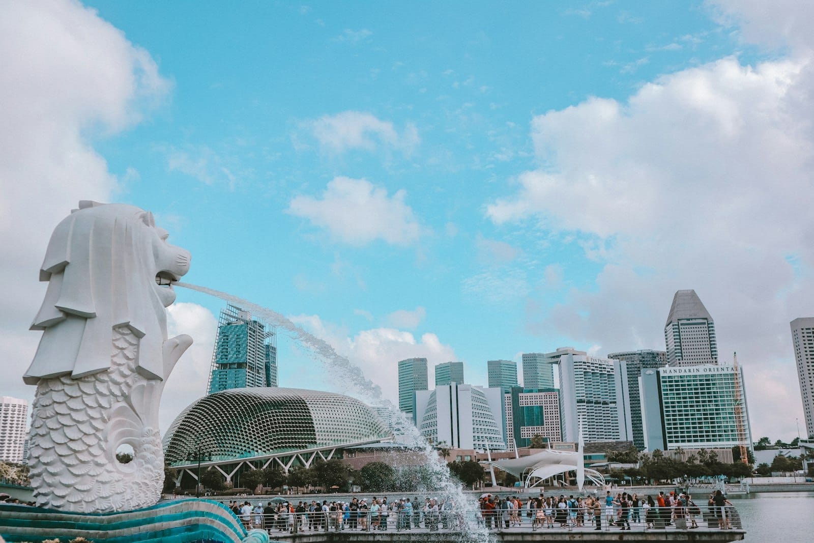 A icônica estátua do Merlion jorrando água com o skyline moderno de Singapura e seus marcos famosos sob um céu claro com algumas nuvens.