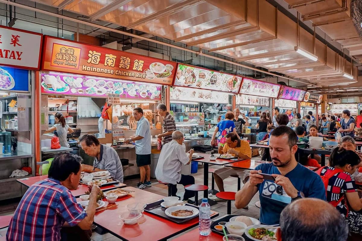 A lively Singapore hawker centre with diners eating at tables and colorful food stalls selling dishes like Hainanese chicken rice.
