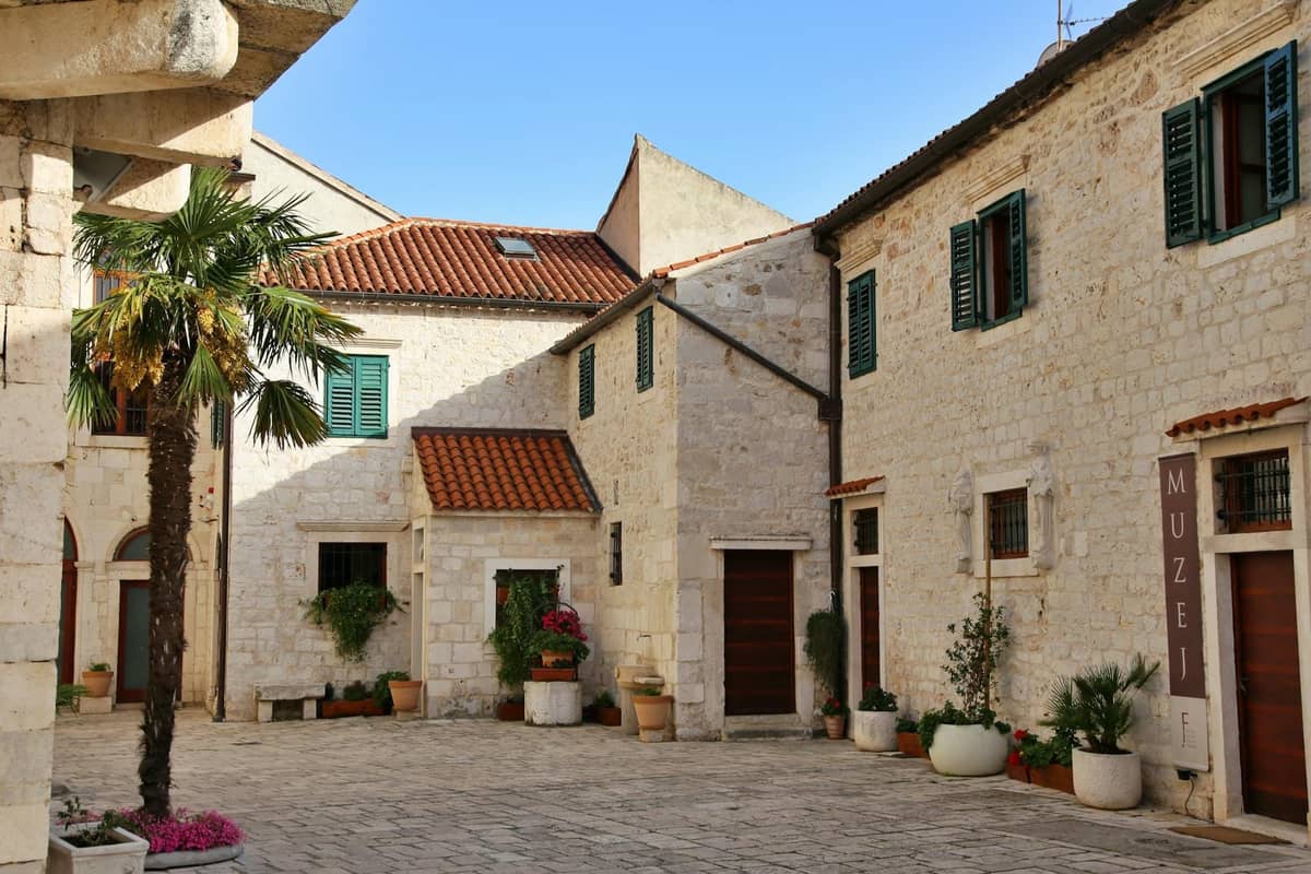Sonniger Steinhof mit Topfpflanzen und Gebäuden, darunter ein sichtbares Schild mit der Aufschrift 'MUZEJ', das auf ein Museum in Split, Kroatien, hinweist.