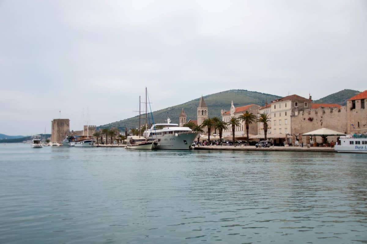 Blick auf die Uferpromenade von Trogir mit historischen Steingebäuden, Palmen und im Hafen liegenden Booten an einem bewölkten Tag.