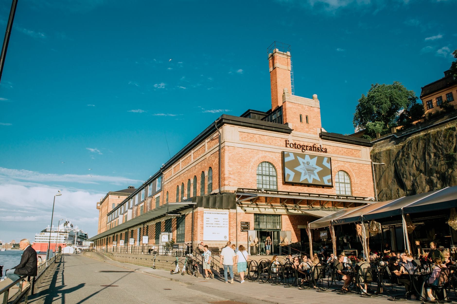 ด้านนอกของพิพิธภัณฑ์ Fotografiska ในสตอกโฮล์มที่มีผนังอิฐ ป้ายบอกทางชัดเจน ที่นั่งกลางแจ้ง และผู้คนเพลิดเพลินกับทางเดินริมน้ำ