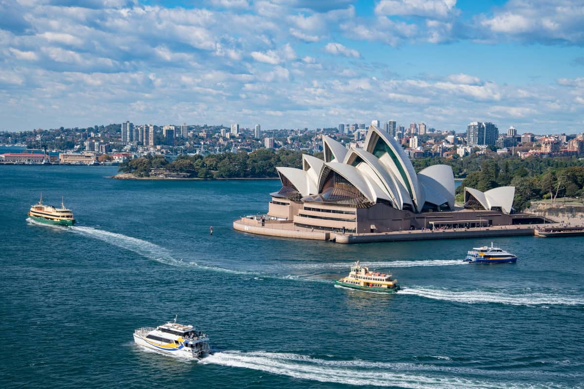 Wide view of Sydney Opera House on the harbor with several ferries crossing the water, and city skyline in the background under a blue sky.