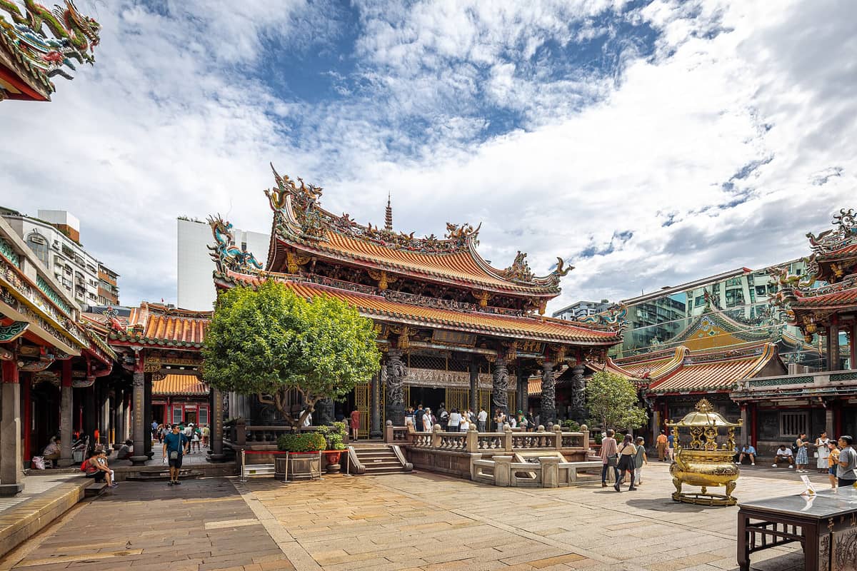 Hofansicht des Longshan-Tempels in Wanhua, Taipeh, mit verzierten Dächern, gemeißelten Säulen, goldenen Details und Menschen, die das historische Tempelgelände erkunden.