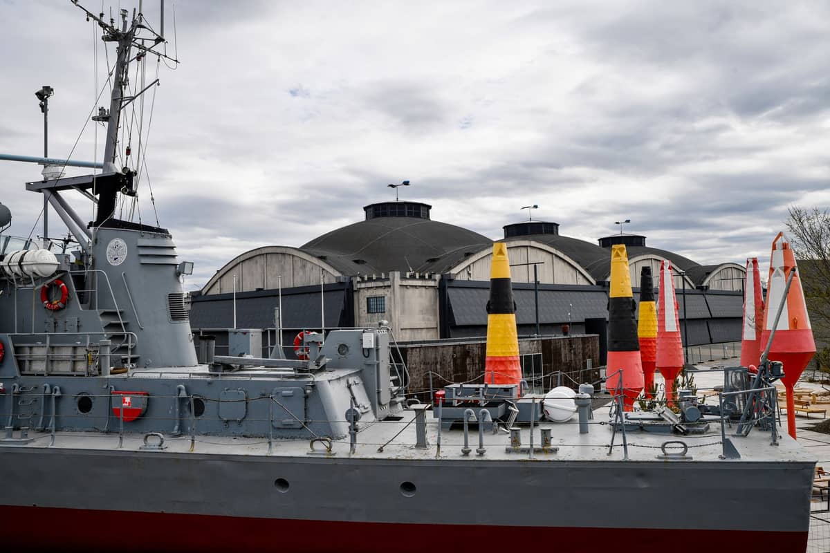 Graues Kriegsschiff vor den markanten Bogenhangar für Wasserflugzeuge des Lennusadam Maritime Museum in Tallinn angedockt, mit bunten Seetonnen an Deck.