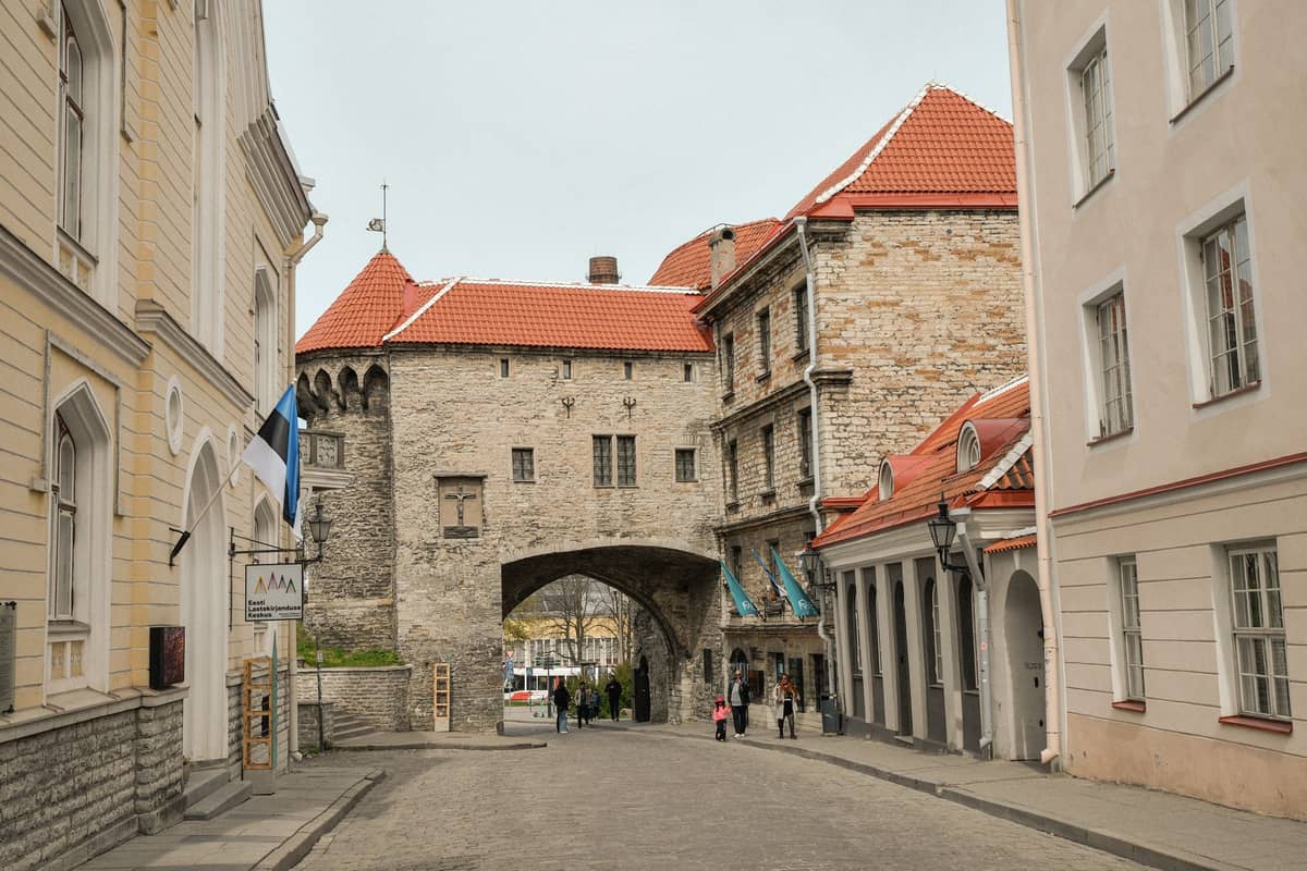 Mittelalterliches Steintor mit rotem Ziegeldach, flankiert von historischen Gebäuden auf einer Kopfsteinpflasterstraße in der Tallinner Altstadt.