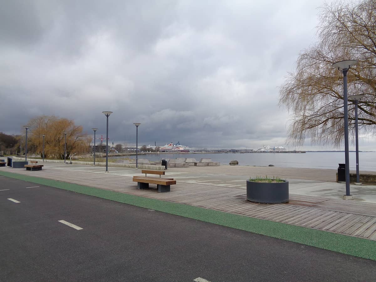 Strandpromenade in Tallinn mit Bänken, Radweg und weitem Blick auf den Hafen und Fähren unter bewölktem Himmel.