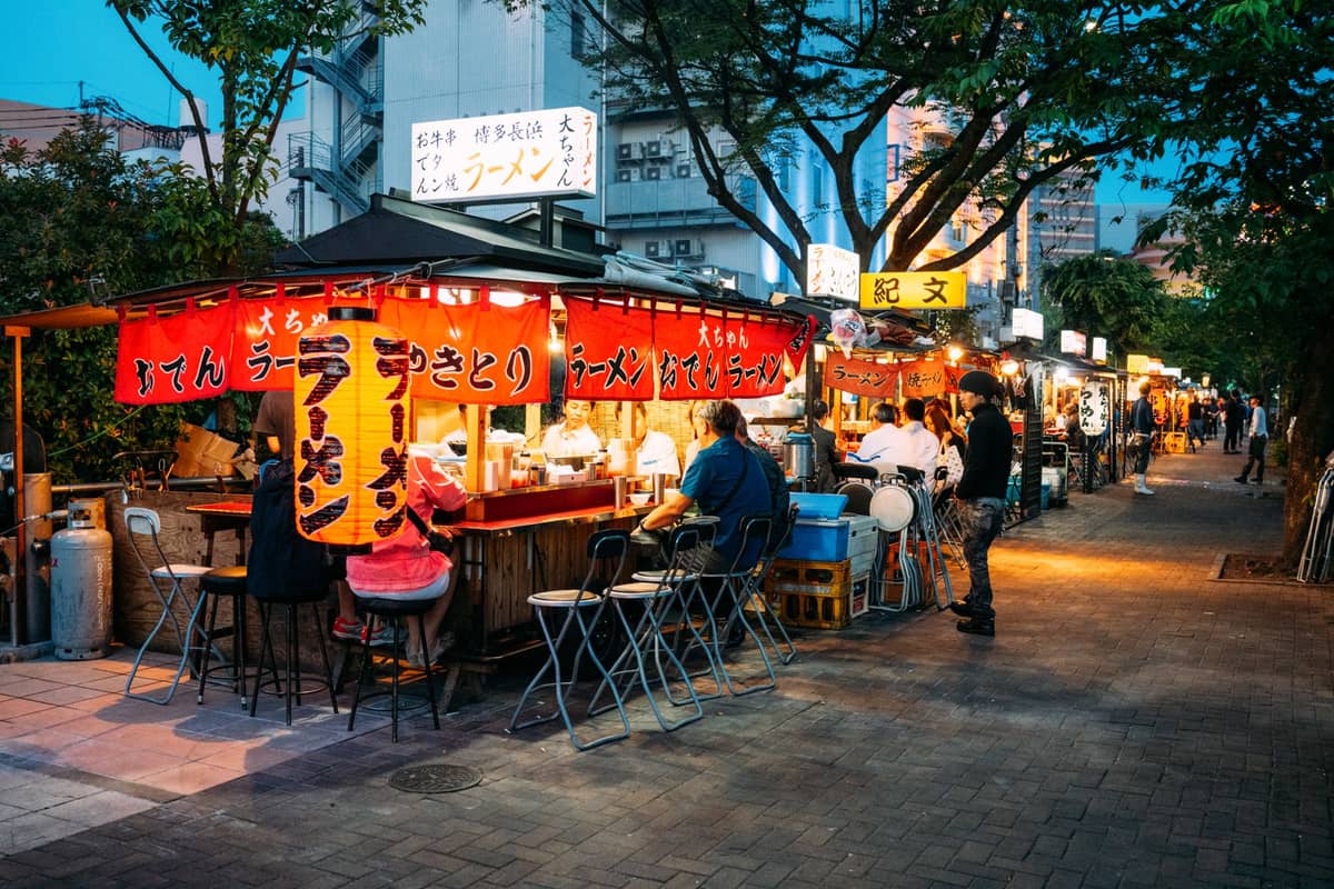 Outdoor ramen food stalls with customers sitting and eating under bright signs on a bustling Tokyo street at dusk.