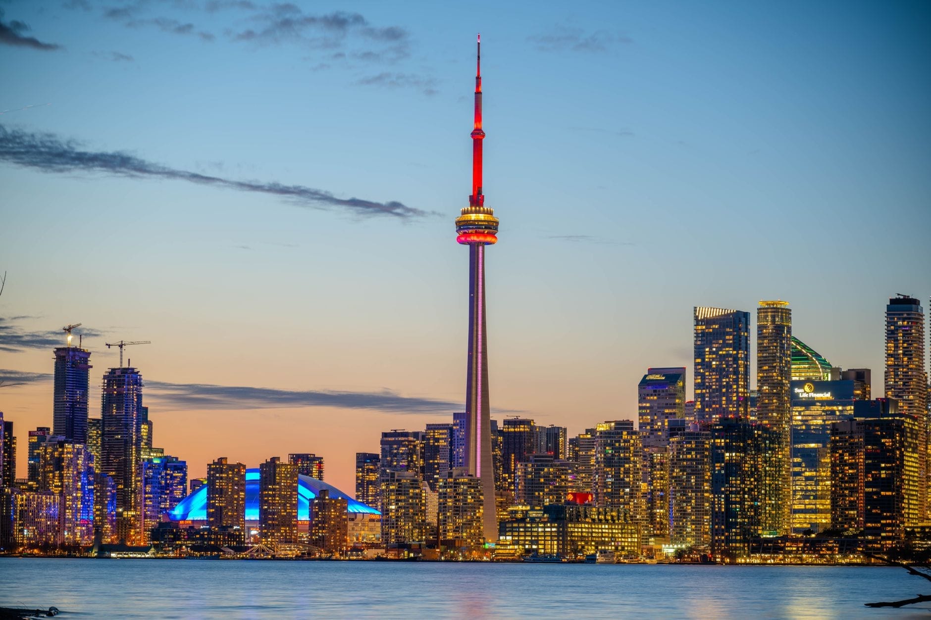 Skyline de Toronto ao entardecer com a CN Tower iluminada, cercada por arranha-céus acesos, vista do Lago Ontário com um céu colorido sobre a cidade.