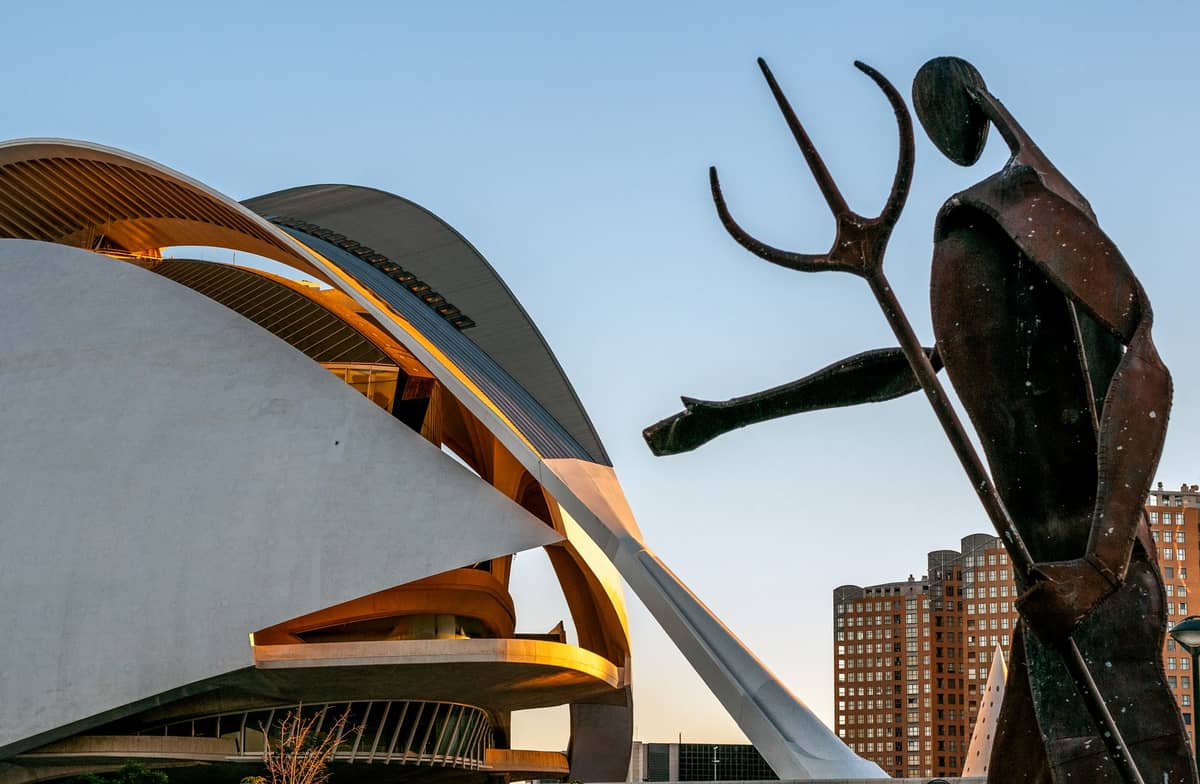 Arquitectura moderna de la Ciudad de las Artes y las Ciencias de Valencia con escultura y edificios residenciales al fondo al atardecer.