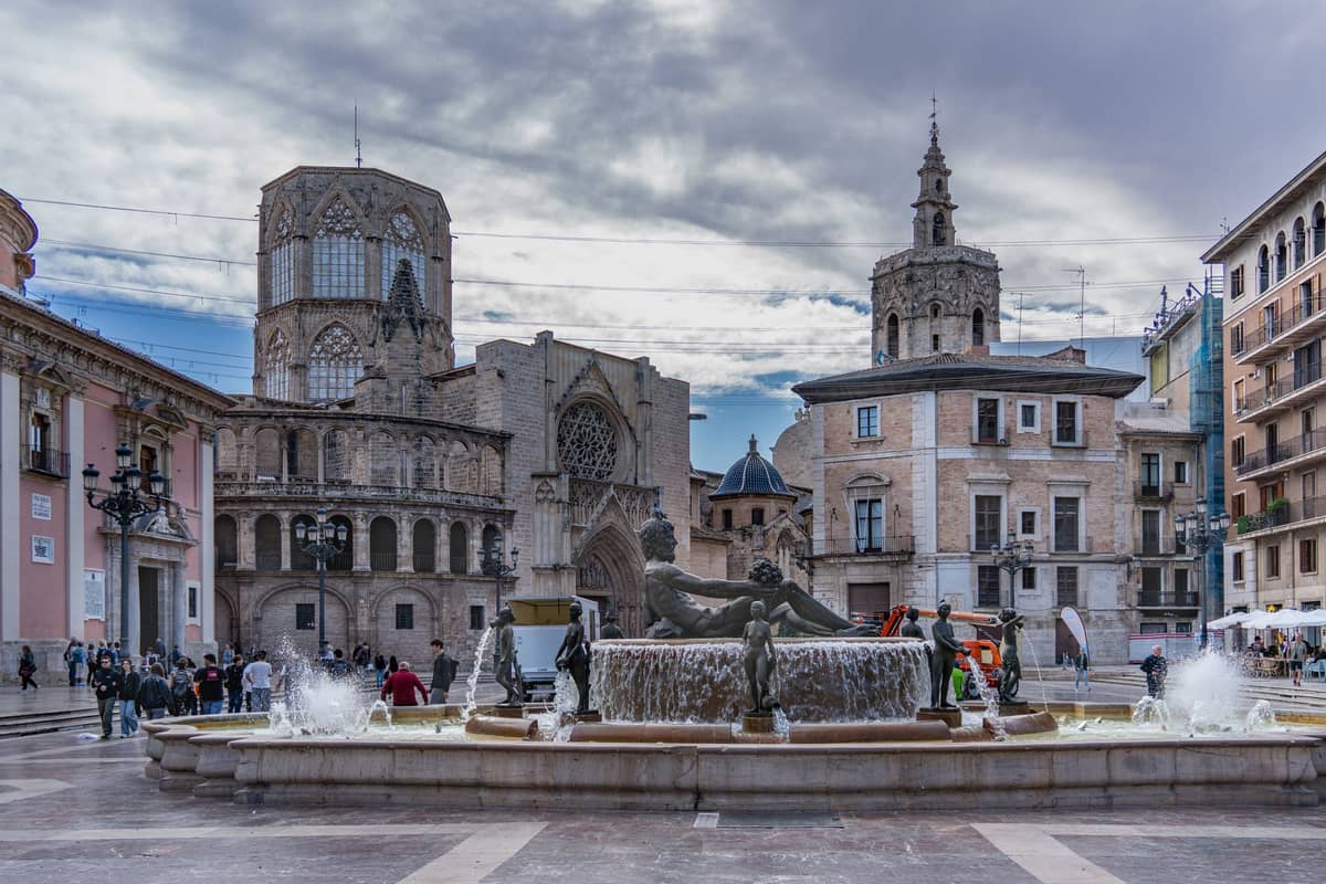 Vista amplia de la Plaza de la Virgen con la Catedral de Valencia, su campanario y una fuente central rodeada de edificios históricos y personas.