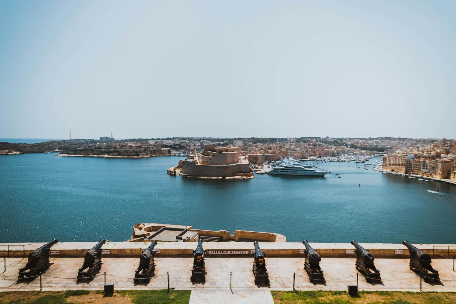 Vista panorâmica do Grand Harbour de Valletta com canhões históricos em primeiro plano e o imponente Forte St. Angelo do outro lado das águas, sob um céu azul e limpo.