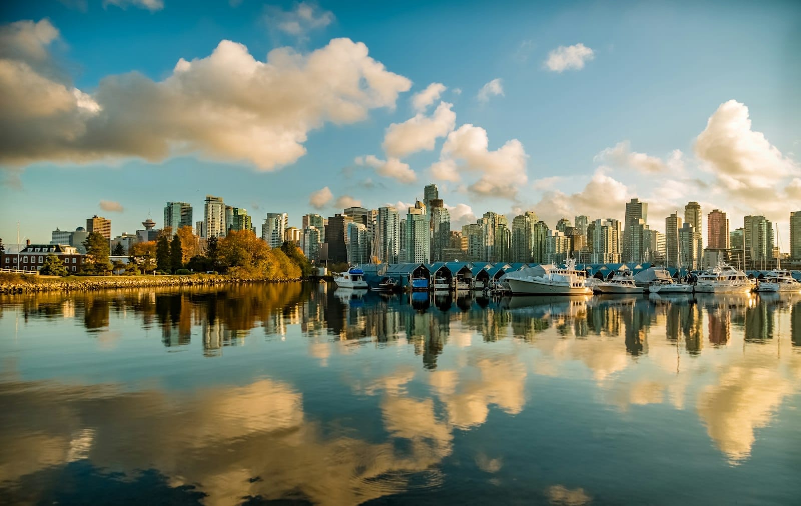 Skyline de Vancouver ao amanhecer com barcos ancorados em águas calmas, reflexos de nuvens e os prédios do centro sob um céu azul.