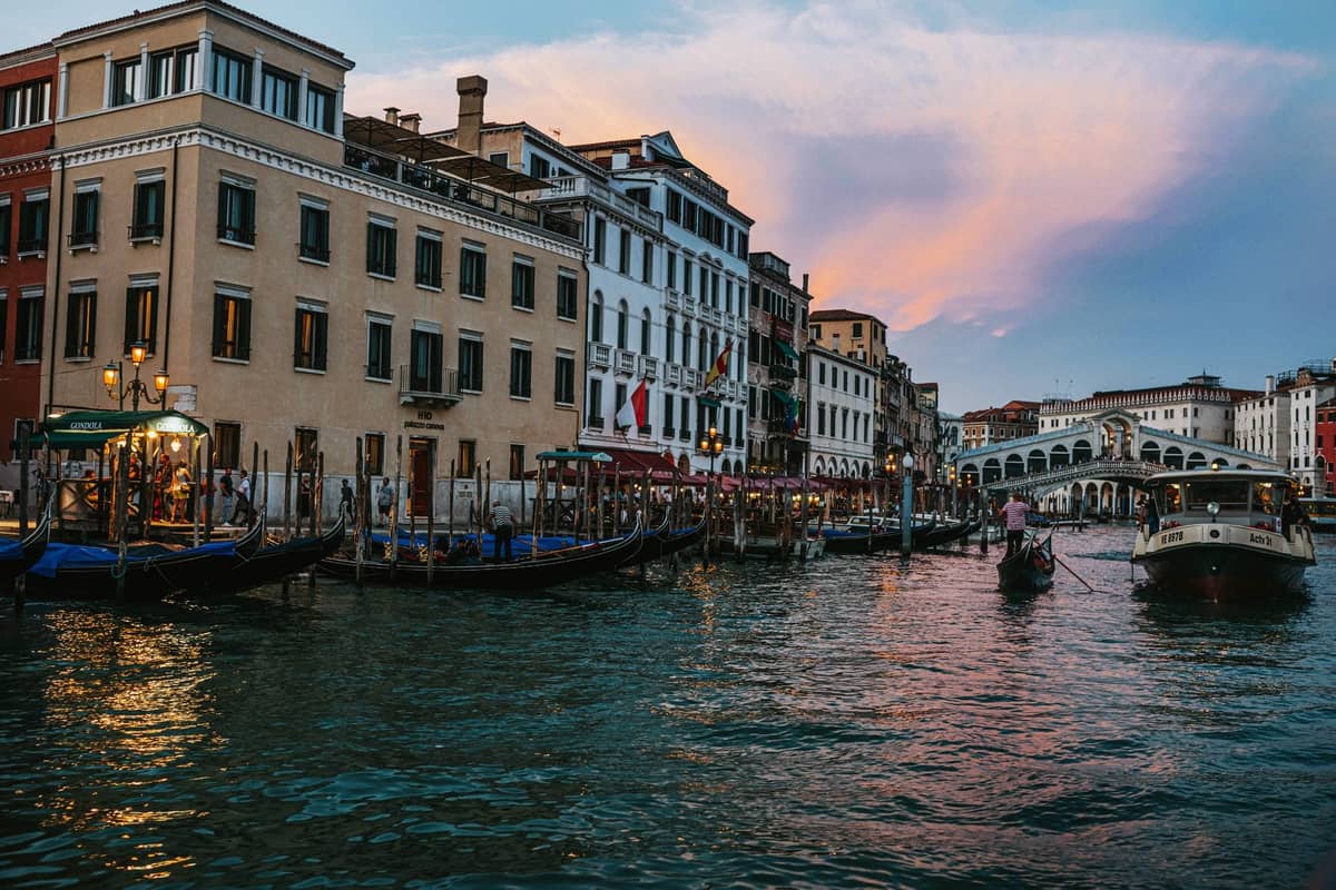 Venedigs Canal Grande bei Sonnenuntergang mit Gondeln, Palazzi am Wasser und der ikonischen Rialtobrücke im Hintergrund.