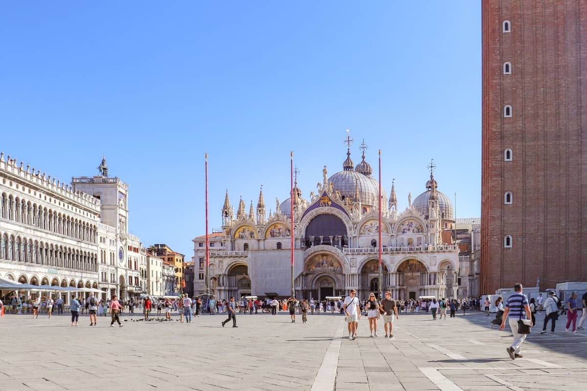 Weitwinkelansicht der Basilika San Marco und des umliegenden Platzes in Venedig mit Spaziergängern, klarem blauem Himmel und einem Teil des Campanile im Bild.