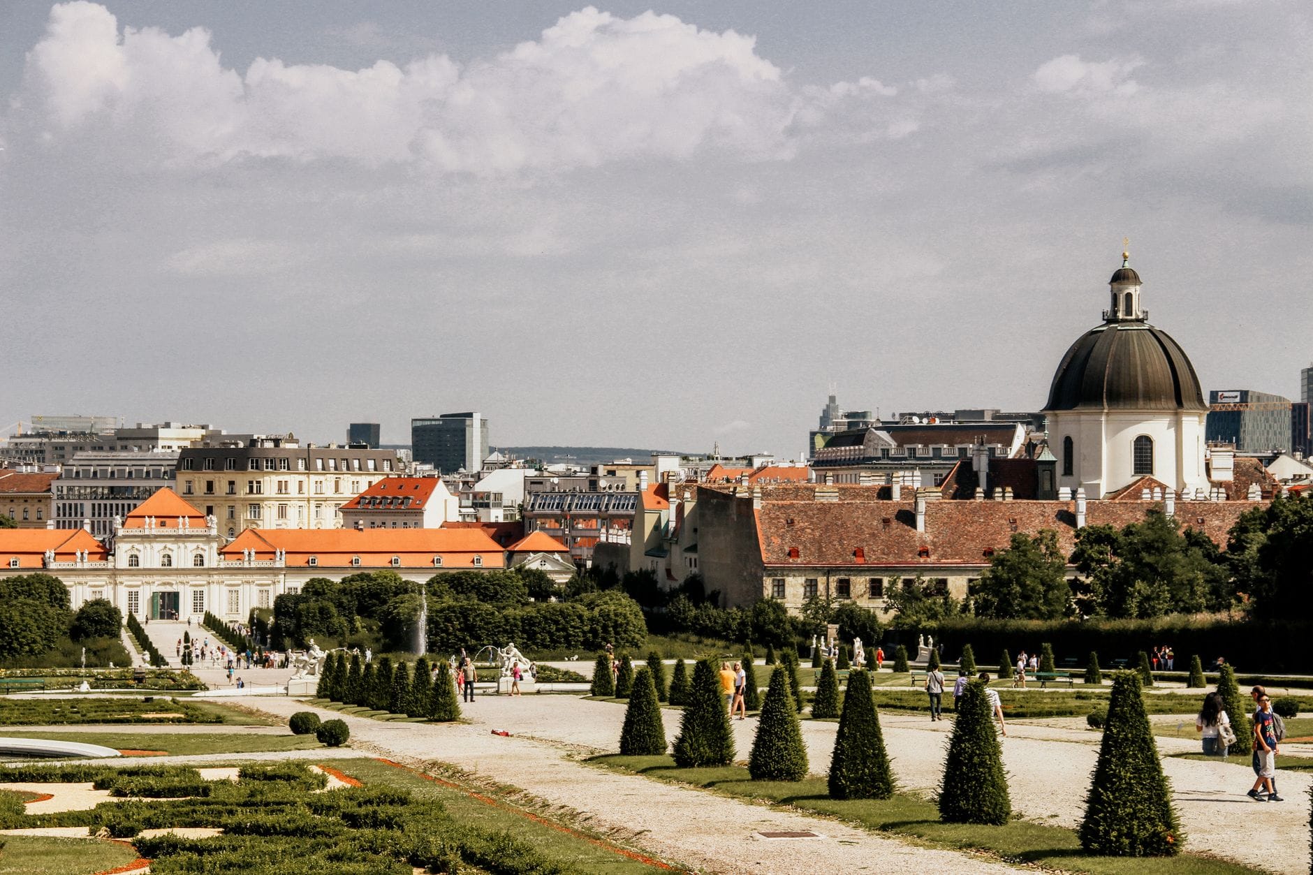 Blick auf die Belvedere-Gärten in Wien mit gepflegten Hecken, spazierenden Menschen, Stadtgebäuden und einer Kirchenkuppel im Hintergrund unter einem teilweise bewölkten Himmel.