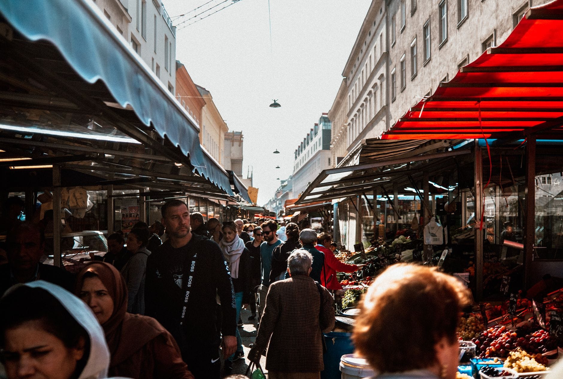 Belebte Außenmarktszene in Wien mit Menschenmassen und bunten Marktständen auf beiden Seiten einer engen Straße.