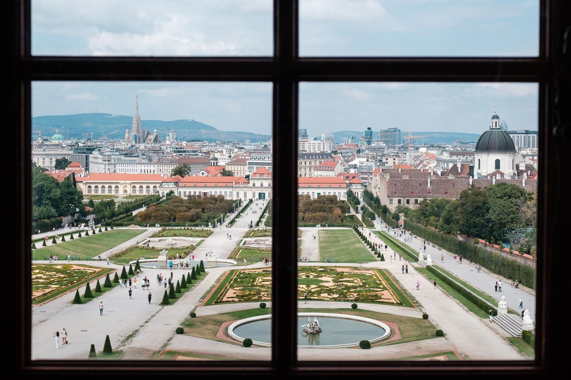 Blick aus einem Fenster auf die symmetrischen Gartenanlagen und die Stadtsilhouette Wiens, mit Spaziergängern auf dem weitläufigen Gelände und historischen Gebäuden im Hintergrund.